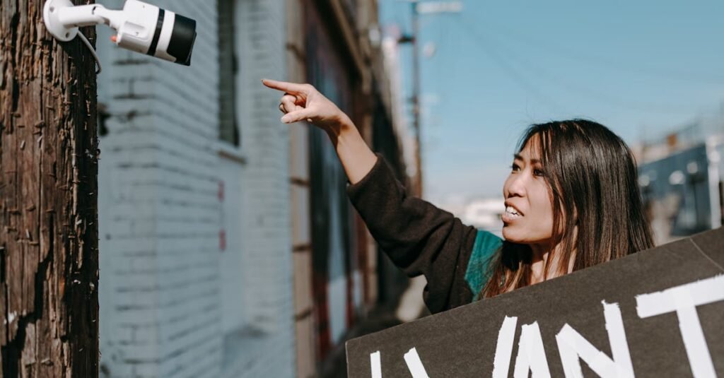 A woman holds a placard, pointing at a CCTV camera during an outdoor protest.
