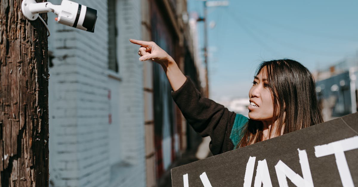 A woman holds a placard, pointing at a CCTV camera during an outdoor protest.